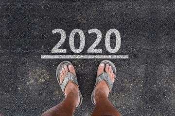 happy new year 2020.. man with dirty feet in slippers is standing on asphalt next to line and 2020 year