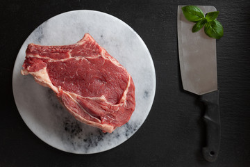Raw fresh Beef steak with bone, spices and seasonings on stone cutting board. Top view, close upon black background