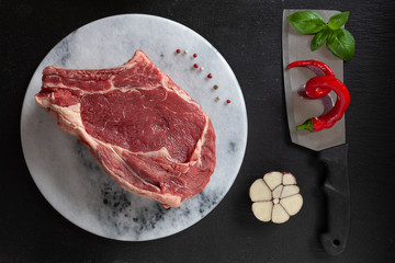 Raw fresh Beef steak with bone, spices and seasonings on stone cutting board. Top view, close upon black background
