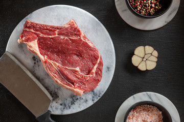 Raw fresh Beef steak with bone, spices and seasonings on stone cutting board. Top view, close upon black background