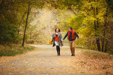 Fototapeta premium Couple weared in autumn-style clothes (oranfe scarf and vest) running through the autumn landscape. Alley covered with yellow foliage. Autumn walk outdoors. Two lovers in autumn park. Romantic dating