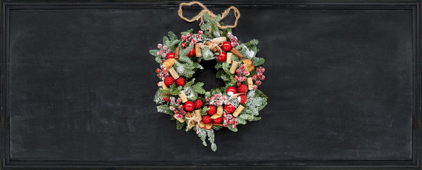 Christmas wreath made of fir branches, dried apples, cinnamon, red berries, bottle caps, red balls hanging on a black chalk board.