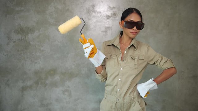 Attractive young girl builder with paint roller in uniform, protective glasses and gloves smiling over concrete wall background