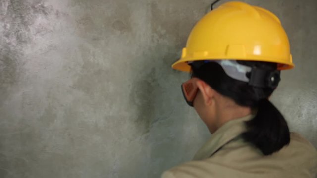 Back view of girl builder with paint roller in uniform, helmet, protective glasses and gloves painitng concrete wall