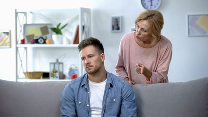 Nervous mother talking to adult son sitting on sofa at home, family conflict