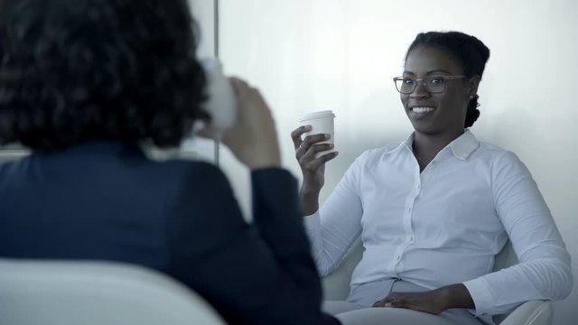 Multiethnic businesswomen drinking from paper cups. Selective focus of cheerful female colleagues in formal wear sitting together and talking in office. Cooperation concept