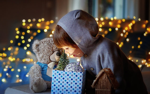 Christmas mood. Cute little excited child with teddy friend looking inside gift box with christmas toys and light from it with garland lights bokeh at background at home.
