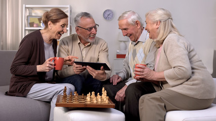 Smiling pensioners watching online video tablet together, playing chess, hobby