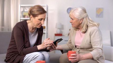 Senior woman using smartphone sitting with mature female friend on sofa, leisure
