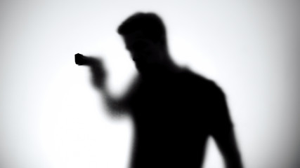 Shadow of male killer aiming gun through glass wall, contract murder, crime