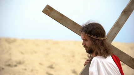Jesus in crown of thorns carrying heavy cross, sacrifice for sinners salvation