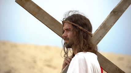 Jesus with bloody forehead and crown of thorns carrying heavy cross, bible event