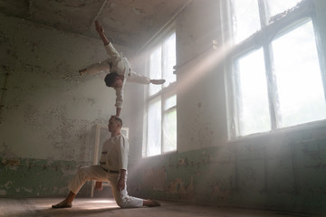 Male duo making acrobatic tricks wearing costume of insane people in abandoned room with sun rays...