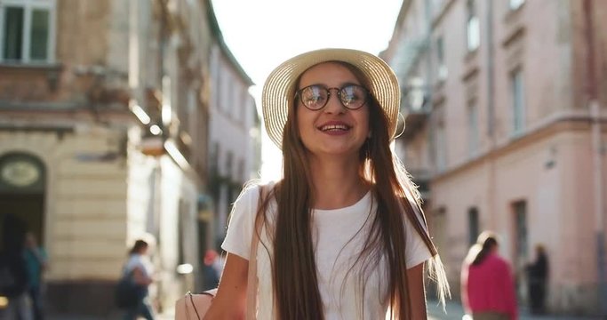 Trendy student in fedora hat, round glasses and hipster backpack. Close up view of Pretty Young Woman Walking like a Model. Looking at the Camera. Wearing Stylish Straw Hat. Having Elegant Make up.
