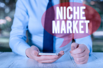 Writing note showing Niche Market. Business concept for Subset of the market on which specific product is focused Female business person sitting by table and holding mobile phone