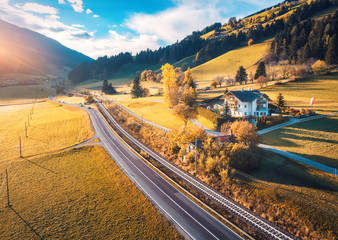 Aerial view of the road in mountain valley at sunset in autumn. Top view of cars on asphalt roadway, house, railroad, hills with orange meadows, blue sky, yellow trees, buildings in fall. Landscape