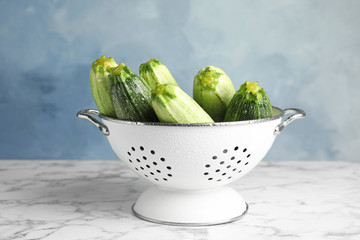 Colander with fresh ripe green zucchinis on marble table against blue background