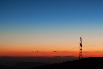 Photograph of a sunset over the mountains in Jordan, with a pole for electricity cables. Beautiful orange, blue, light blue and yellow colors.
