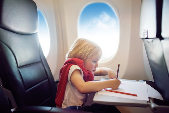 Charming kid traveling by an airplane. Joyful little boy sitting by aircraft window during the flight. Child drawing picture.