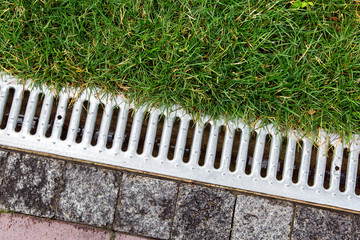 iron grate of a storm drainage system on the side of a footpath made of pavers near a green lawn top view with rain water.