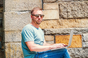 Fototapeta na wymiar Young American Man with beard, traveling, working in New York City, wearing light green T shirt, blue jeans, sunglass, sitting outside on school campus under sun in summer, working on laptop computer