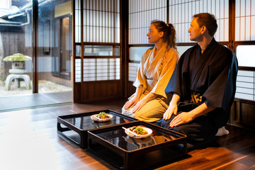 Couple in kimono seiza sitting at traditional Japanese home ryokan room by table plates by shoji sliding paper doors, looking at peaceful garden with stone lantern