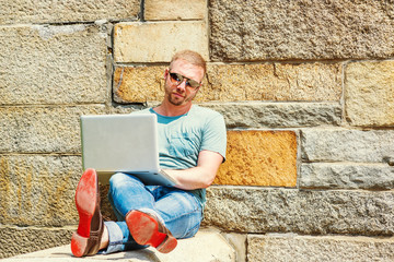 Fototapeta premium Young American Man with beard, traveling, working in New York City, wearing light green T shirt, blue jeans, sunglass, sitting outside on school campus against rocky wall, working on laptop computer..