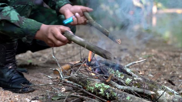  4K Medium close up shot of military soldier making campfire from tree branch in forest near stream brook. Burning flames firewood campfire in forest woodland camping. 
