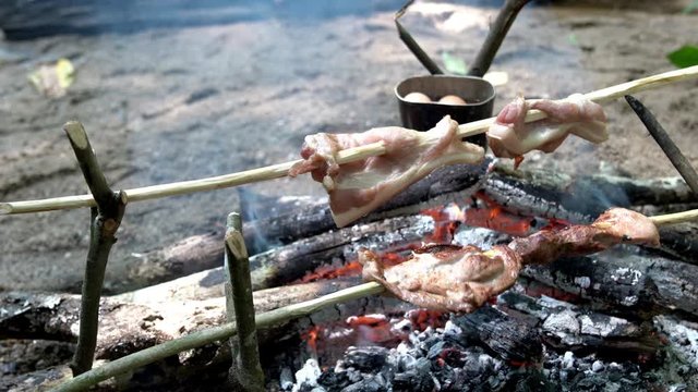 4K Wide shot of grilled BBQ raw pork red meat with boiling egg in camping pot on bonfire at camping. Cooking roast steak meat on fire flames with smoke at hiking camping in forest.