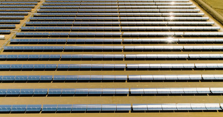 Solar panels in a solar farm with a factory in the middle. There is the reflection of the sun in the the panels which produce renewable energy, solar energy - aerial view with a drone - environment an