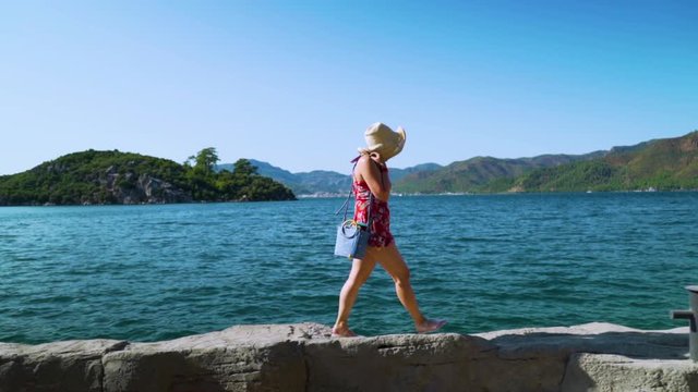 This shot depicts a panoramic view of a lady in a hat and jelly shoes walking by the sea side in Turkey, Marmaris.