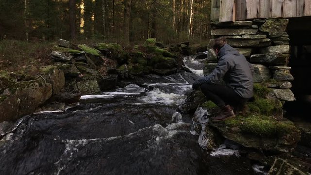 Young man sitting by a small creek running through the woods near Halland, Sweden, during winter time