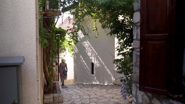 The shot depicts a narrow cobblestone street surrounded by quaint old houses in Marmaris, Tukey. The woman with a straw hat, wearing a floral dress is walking away.