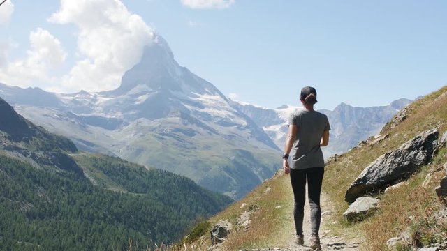 Slow motion athletic woman walking and hiking Swiss Alps Matterhorn trail on dirt path in summer with green grass and mountain range in background