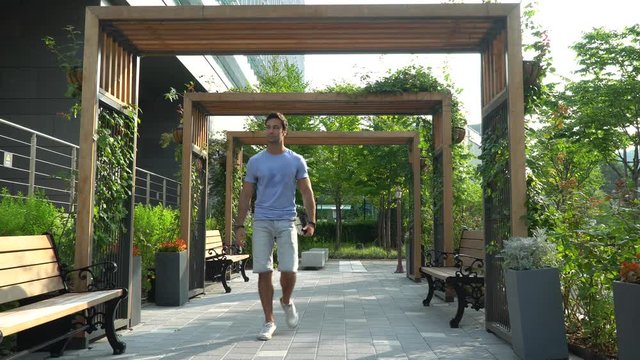 A slightly sweaty, handsome, dark haired, athletic, well dressed European man in shorts and light blue t shirt walks along an elegant tree lined pathway passing under modern wooden structures in Asia