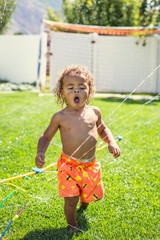 Cute little boy drinking from the sprinklers in the backyard on a hot summertime day. African American boy playing in the outdoors trying to stay cool on a warm summer day
