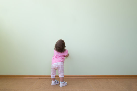 Adorable baby girl in a pink blouse and striped pants stands in front of a green wall and looks up