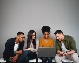Group of diverse friends sitting on chairs in queue watching videos online or using social media app on laptop