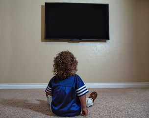 Rear view of a young child watching Television while sitting on the floor of his home. Selective focus on the back of the curly-haired diverse little boy watching sports on TV
