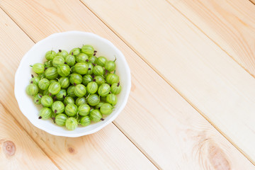 Green berries of fresh gooseberry in white bowl on wooden table with copy space. Summer harvest.