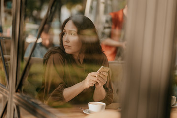 Worried brunette woman with long curly hair sitting at the window in cafe with mobile phone in hands