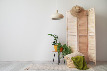 Wooden folding screen with plants on table and wicker basket near light wall in living room - obrazy, fototapety, plakaty