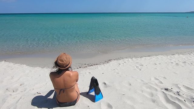 Young beautiful woman sitting on the shore with snorkeling fins resting on the sand observes the horizon with the blue sky and the crystal clear Caribbean sea of Budoni in Sardinia