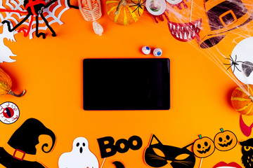 Tablet with blank screen on orange background. Little pumpkins, photo props, masks, spiderwebs, decor for celebration are scattered in circle of canvas. Party accessories. Happy halloween concept.