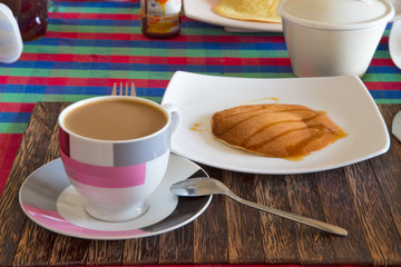  A Cup of coffee with milk and a plate with a pancake is on the table.Horizontally.