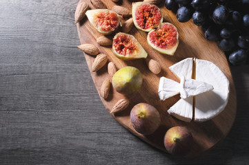 Still life cheese plate on a wooden cutting board lies sliced cheese with mold almonds in the shell and sliced figs next to a bunch of black grapes. Delicious and healthy food concept