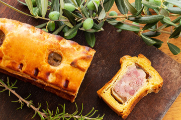 Top view of a pate en croute or pâté en croûte, with rosemary twig and green olives on branch with leaves over a dark wooden cutting board background. French traditional appetiser.