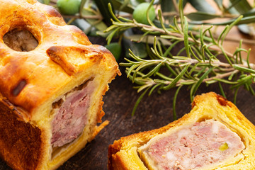 Pate en croute or pâté en croûte or meat pie, with rosemary twig and green olives on branch with leaves over a dark wooden cutting board and a used oak wood background. French traditional appetiser.