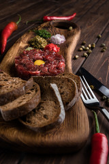 Fototapeta na wymiar Beef Tartar with a raw egg yolk, capers, rye bread and chilis on a wooden board with spoon and fork. Shot made from above on a dark rustic texture