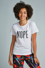 Three-quarter shot of an afro-american girl with curly hair, posing on a grey background. She is wearing white t-shirt with lettering "Nope" and black trousers with floral print. 